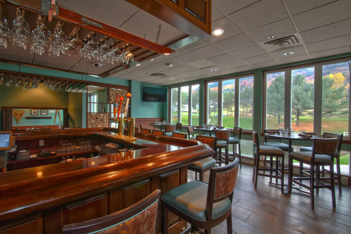 A relaxed bar area with tables and chairs set near large windows, along with hanging glasses and ample space for group dining or casual socializing. The atmosphere is lively yet comfortable, ideal for enjoying meals and drinks together with friends or family.
