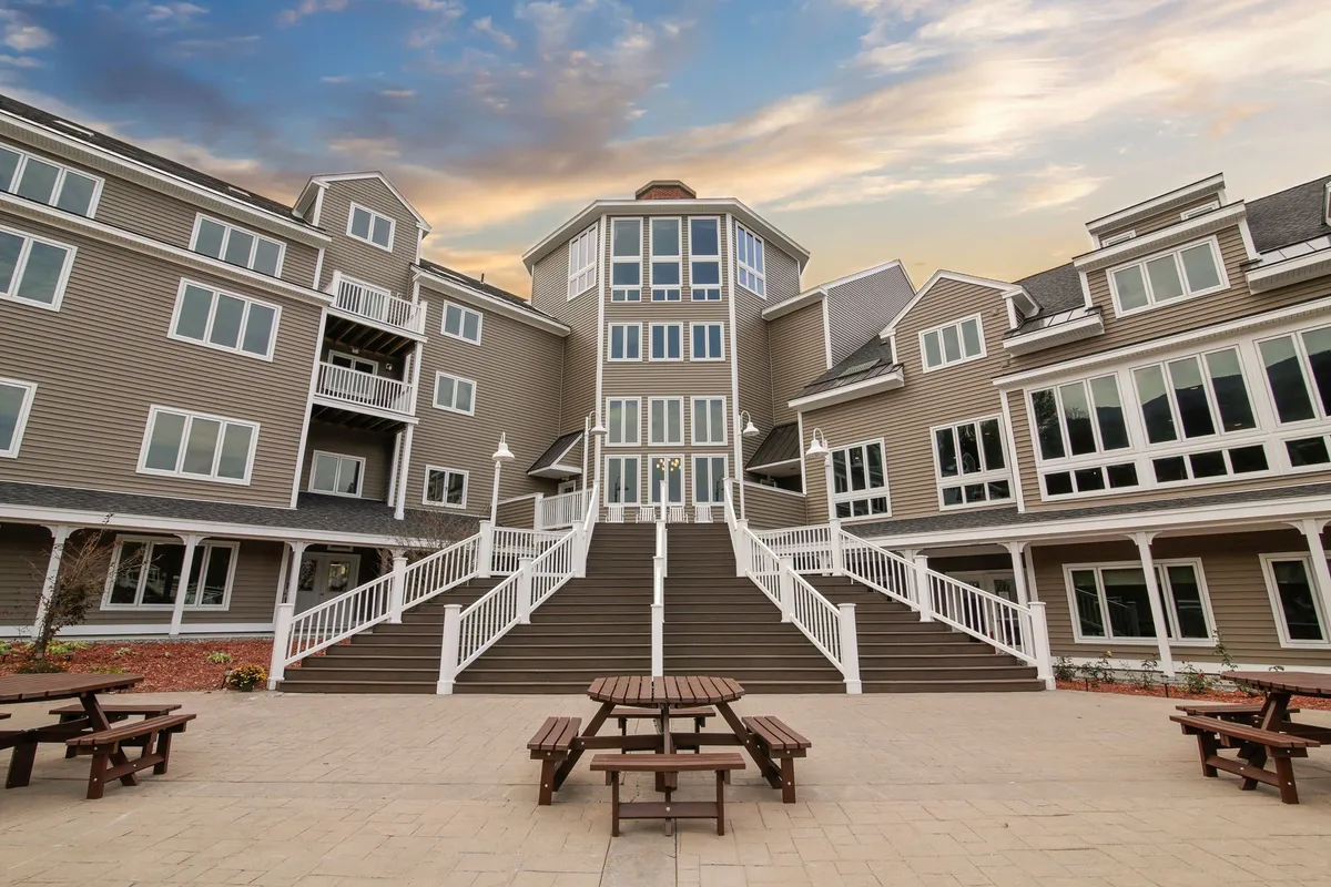 This image captures the property's grand entrance with a sweeping staircase and large windows, set against a tranquil mountain backdrop. Picnic tables are arranged near the entrance, providing spaces for guests to gather and enjoy the fresh mountain air. The scene is bathed in soft, welcoming light, hinting at sunset views from the upper floors and a serene, communal atmosphere.