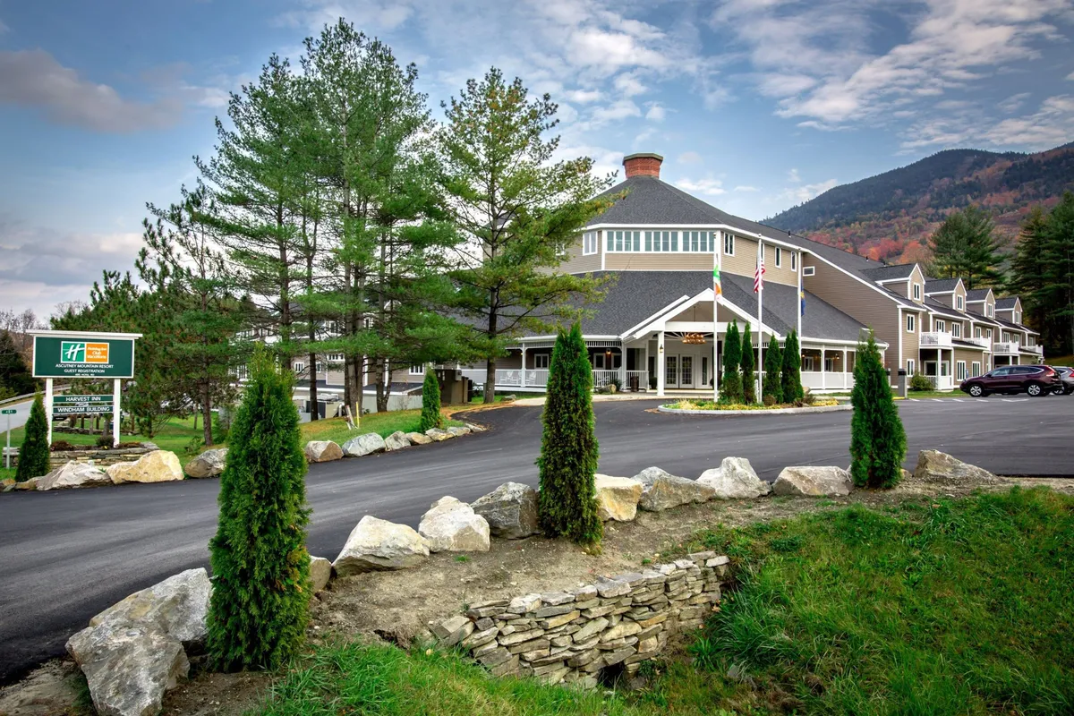 The image features the welcoming hotel entrance framed by beautifully landscaped pathways and flag poles, all set against a stunning mountain backdrop. The scene suggests easy access to nature, with the entrance inviting guests toward both adventure and comfort.