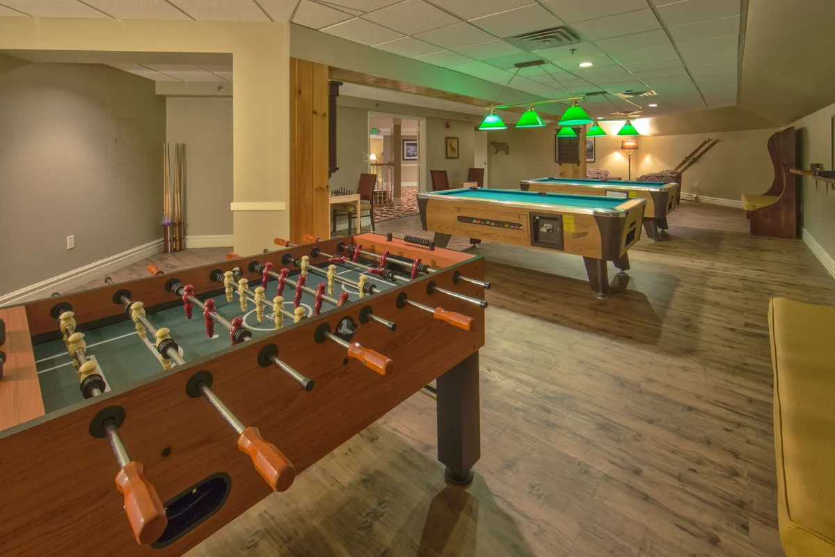 A vibrant recreation room featuring both a foosball and a pool table, complemented by comfortable seating and distinctive green pendant lighting. The area is set up for friendly games, laughter, and casual relaxation, all in a warm, inviting atmosphere.