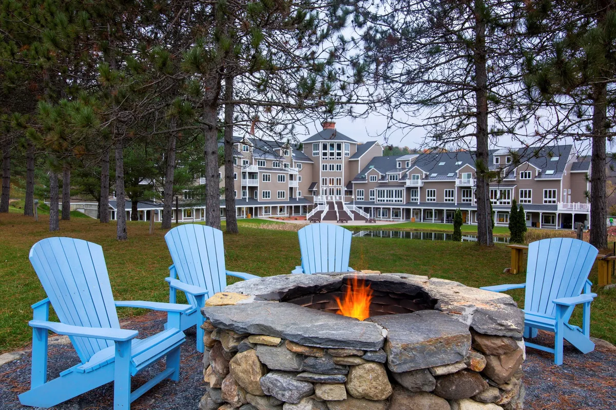 Set outdoors, this image features a cozy fire pit surrounded by blue Adirondack chairs, with the hotel's main building visible in the background. The scene exudes a sense of warmth and community, ideal for relaxing with friends after a day of wellness activities. The backdrop of the mountain setting and hints of sunset lighting contribute to the peaceful mood.