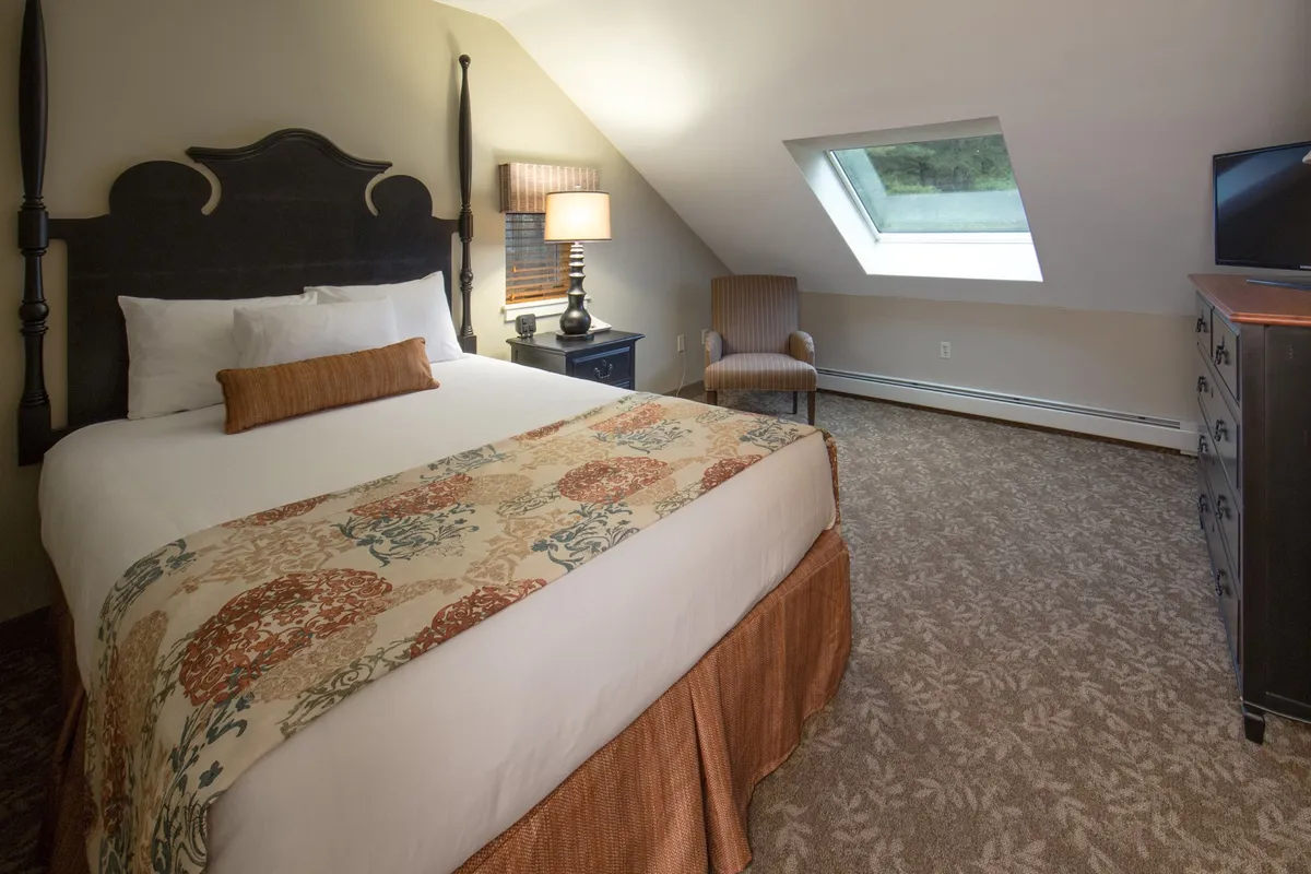 A bright, tranquil bedroom featuring a plush bed adorned with a decorative quilt, a cozy armchair in the corner, a nightstand with a lamp, and a skylight that fills the room with natural light. The space feels calm, inviting, and perfect for unwinding after a long day.